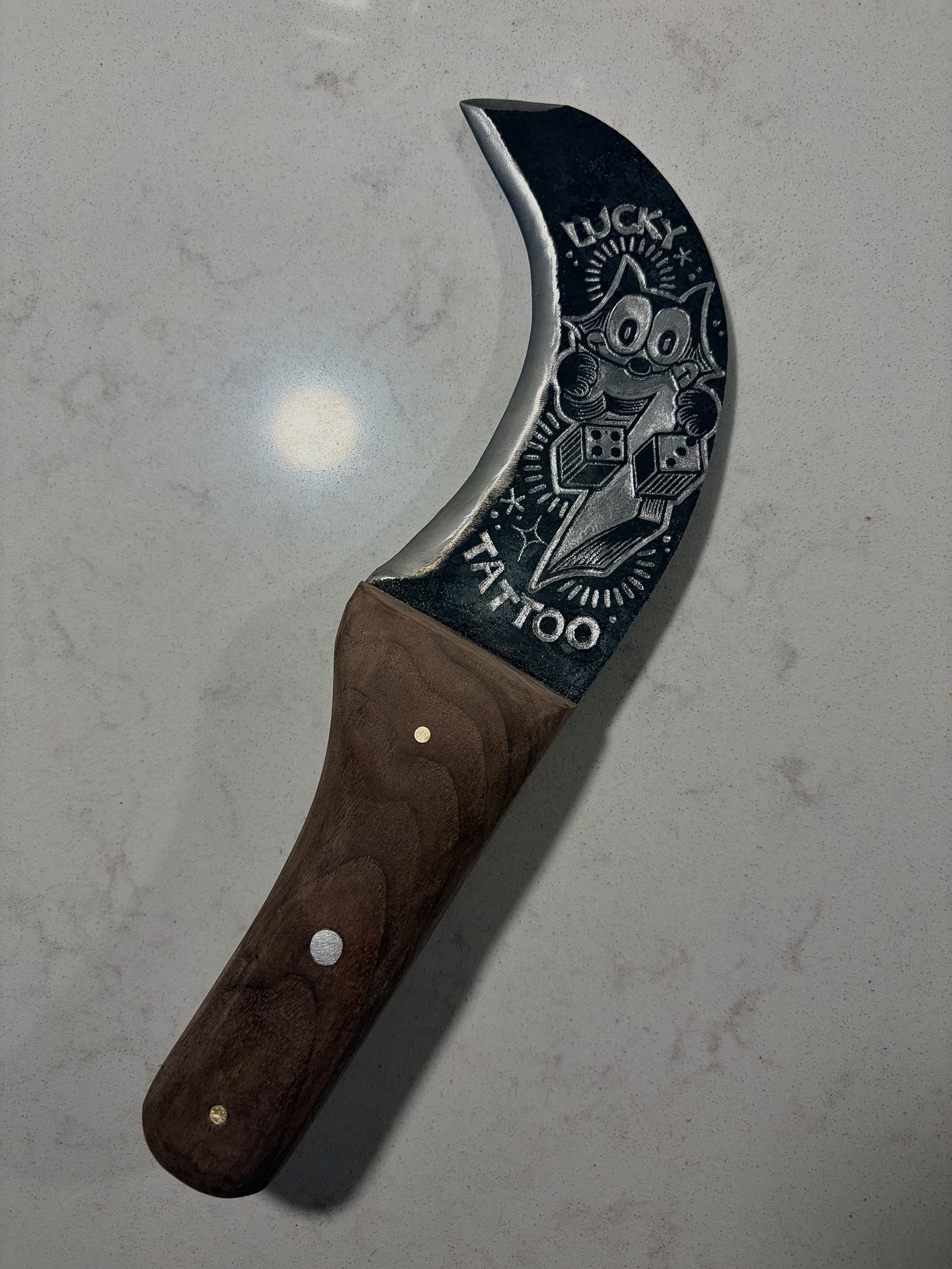 Hand-forged knife with a dark, crescent-curved blade showing a “Lucky Tattoo” design of a cartoon cat holding dice, and a wooden handle with brass and steel pins.