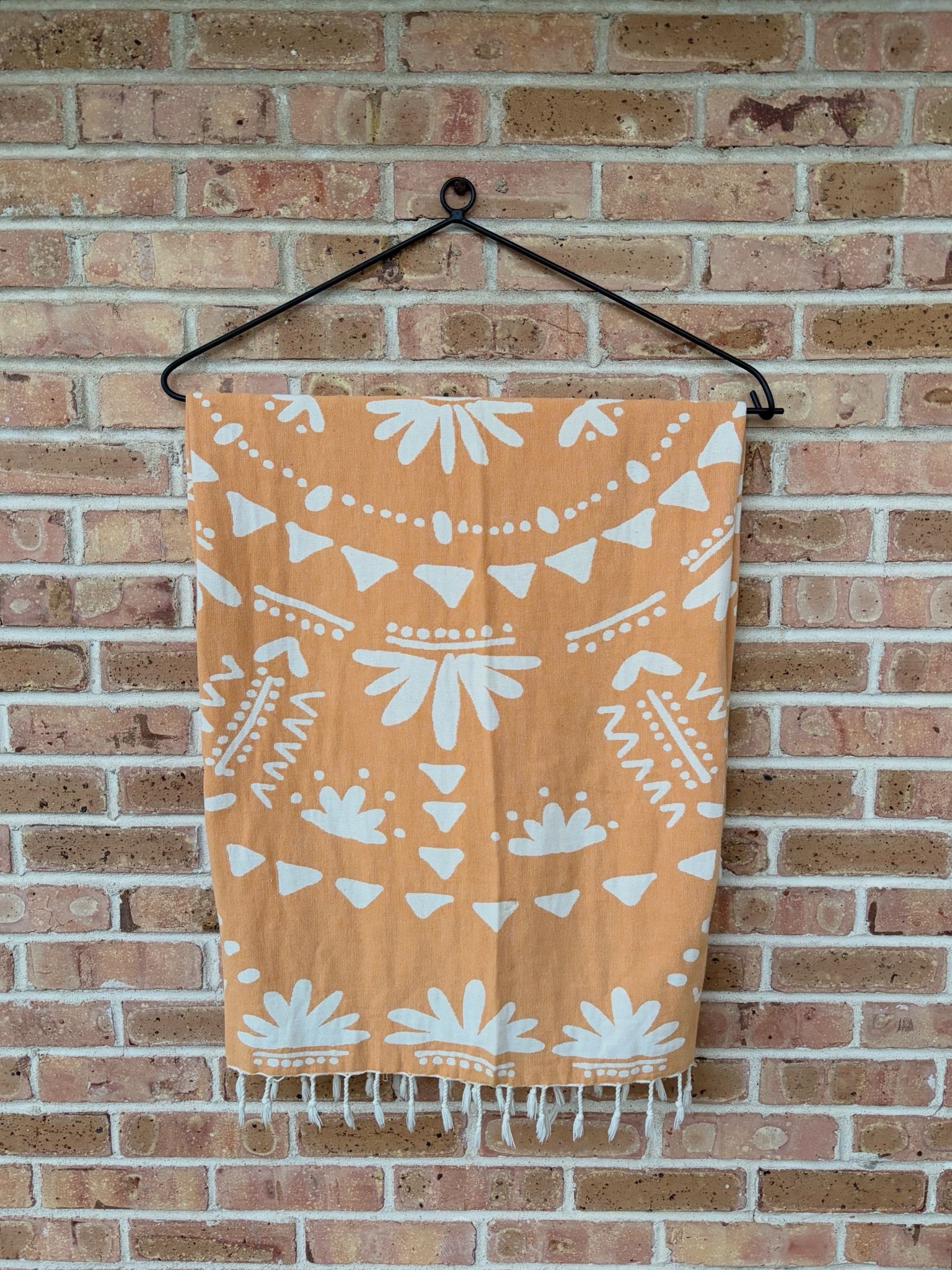 Black steel quilt hanger on a brick wall, holding an orange and cream patterned throw with fringe.