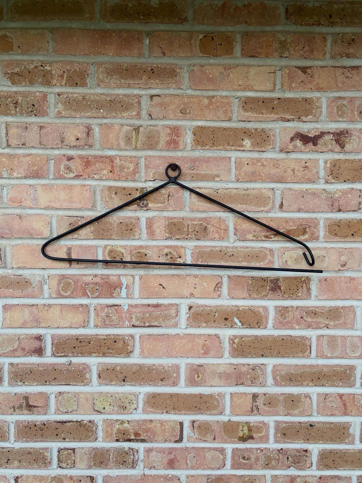 The empty quilt hanger on brick, showing the hanging ring and the open hooked end.