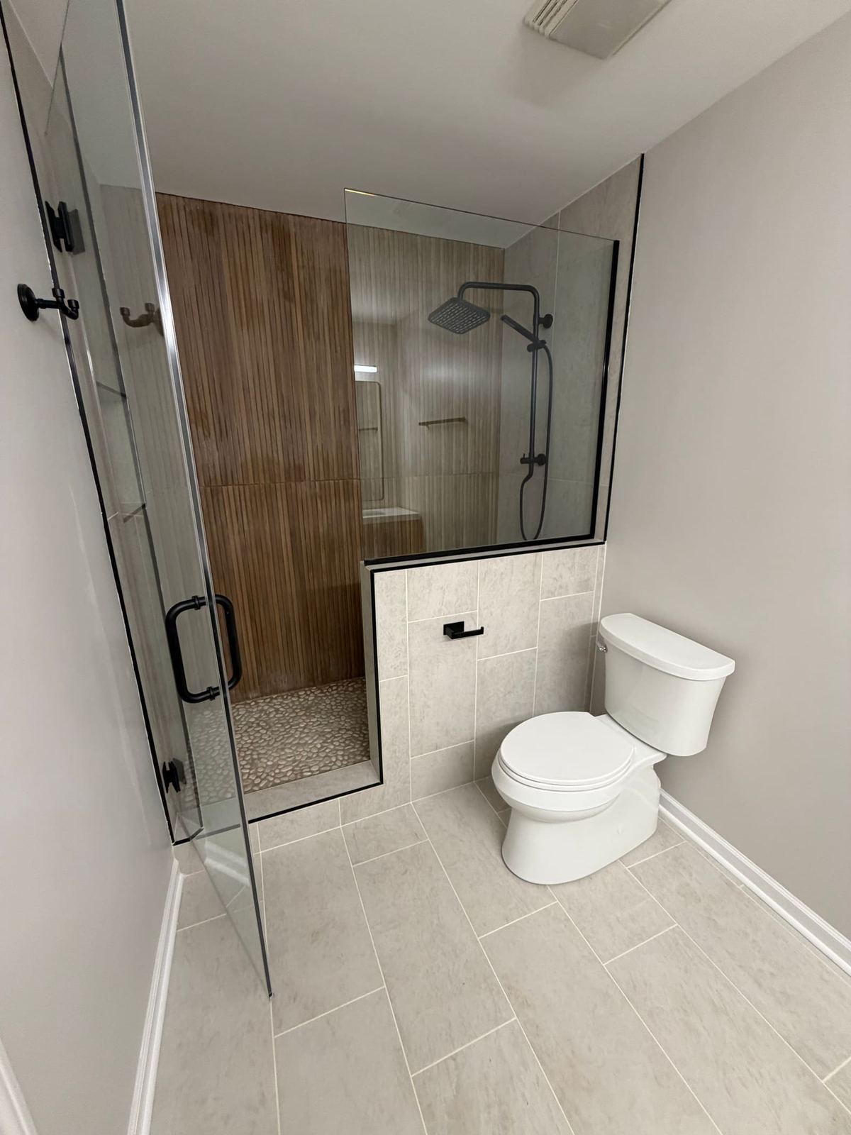 Primary bathroom with a frameless glass shower, fluted wood-tone back wall, pebble floor and black fixtures.